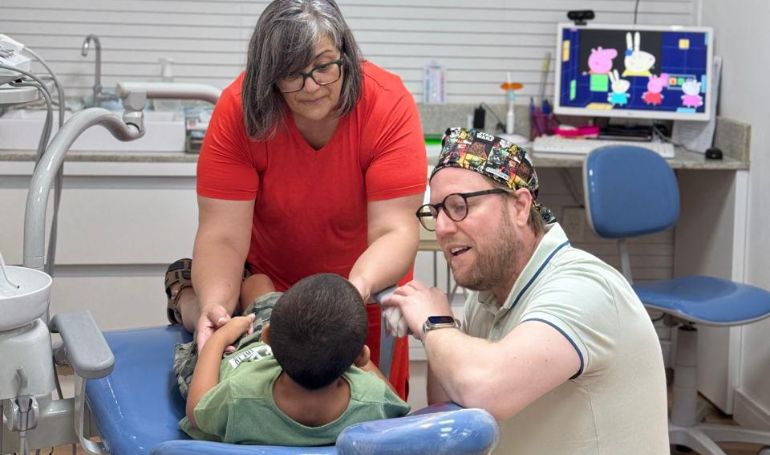 Atendimento odontológico infantil com acompanhante e profissional ao lado da cadeira.